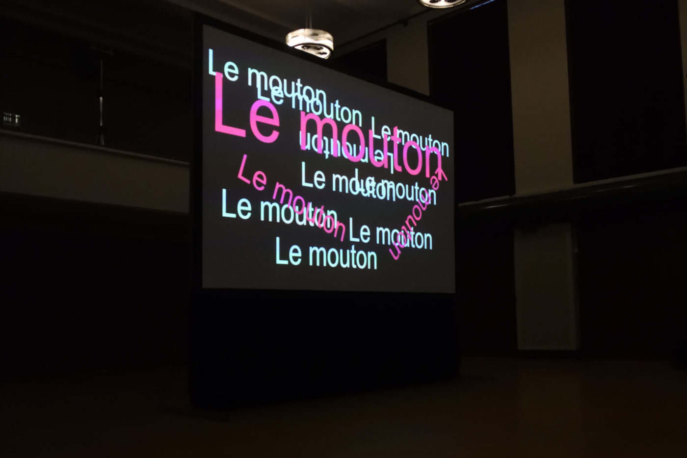 On voit un écran où est projeté plusieurs fois le mot mouton. Le mot est projeté en rose et en blanc, en différentes tailles.