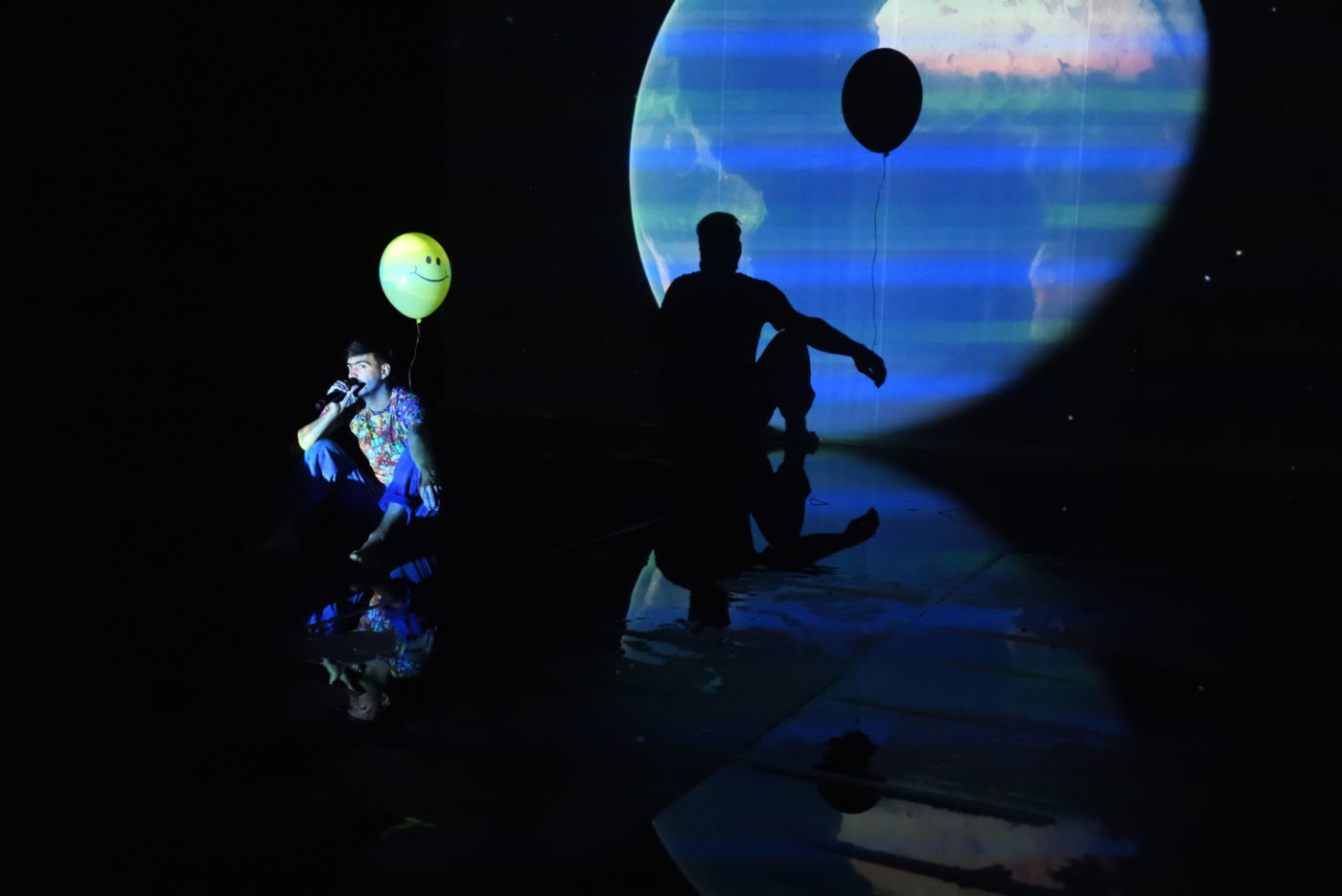 L'artiste Savino Caruso tient un ballon de baudruche dans le dos. Son ombre se reflète en arrière plan dans une projection de la planète Terre. Il parle dans un micro.
