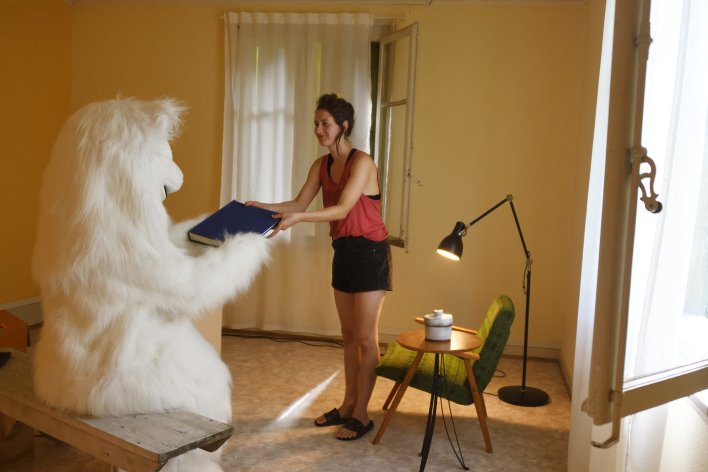 L'artiste Camilla Parini se trouve debout dans une petite pièce. Elle tend un grand livre à une personne déguisée en ours blanc.