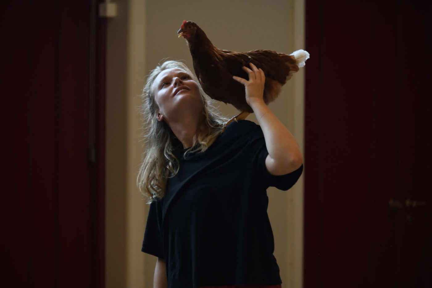 On voit l'artiste Danae Dario porter une poule sur son épaule gauche. Elle le regarde.