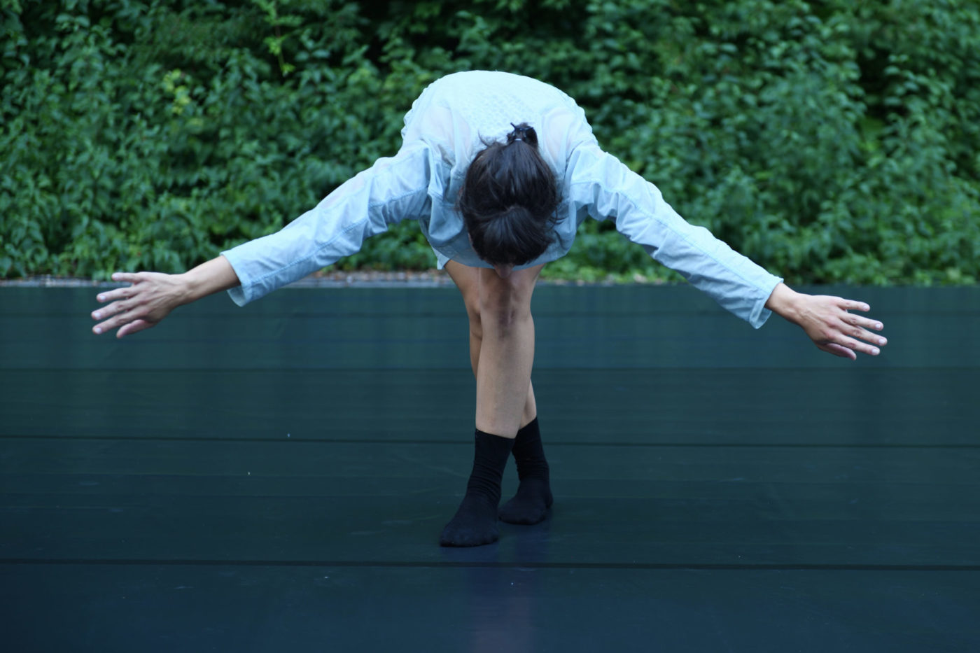 La danseuse Marion Baeriswyl se tient à l'extérieur sur une scène dans une forêt. Elle tire une révérence le haut du corps vers le bas, les bras écartés.