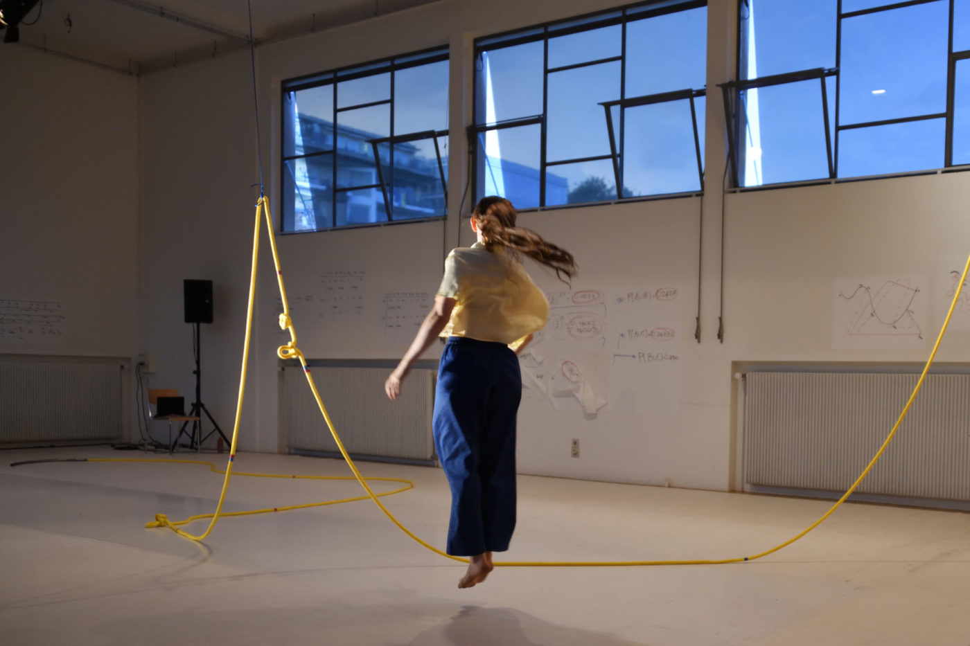 La danseuse Bianca Berger saute à la corde de manière latérale. La grande corde jaune est fixée au bout de deux autres cordes fixées au plafond.