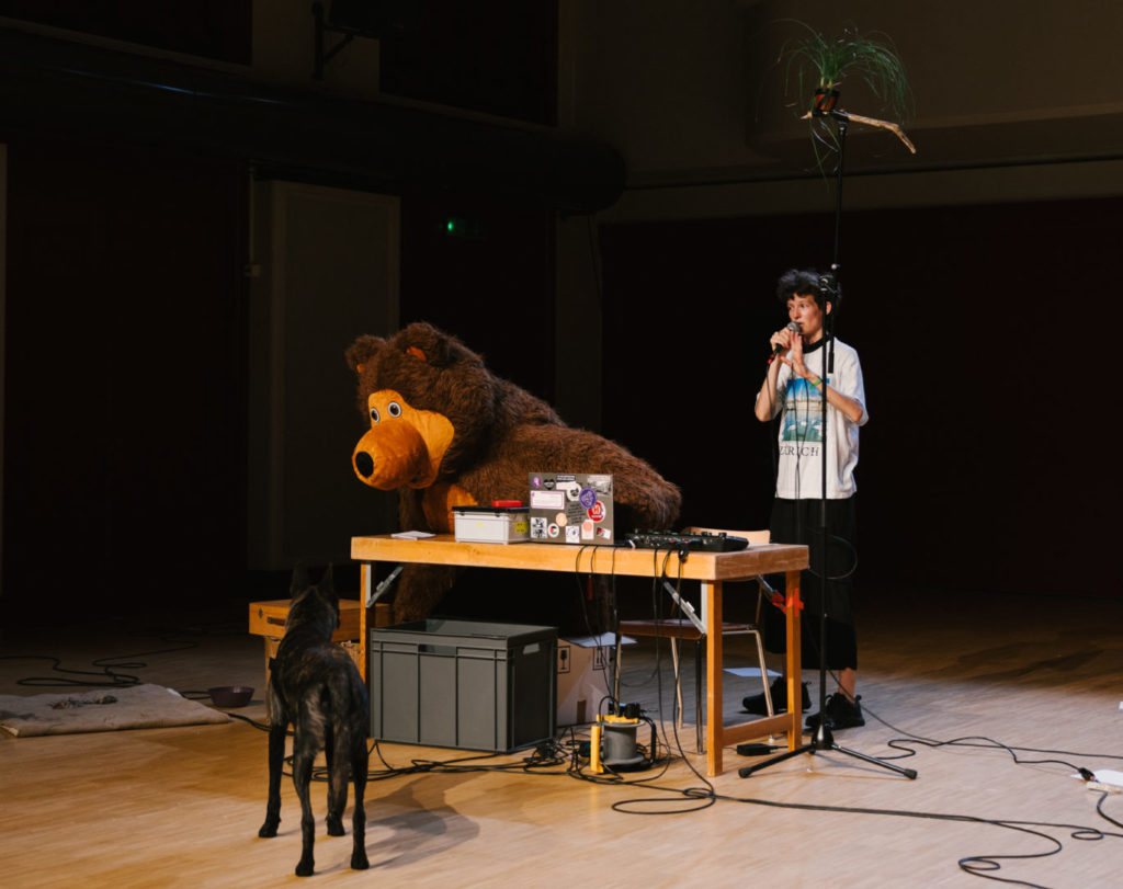 The comedian Elise Perrin stands on the scene with her dog. Both are looking at a big inflatable teddy bear that sits on a table. There is also a plant at the extremity of a micro stand.