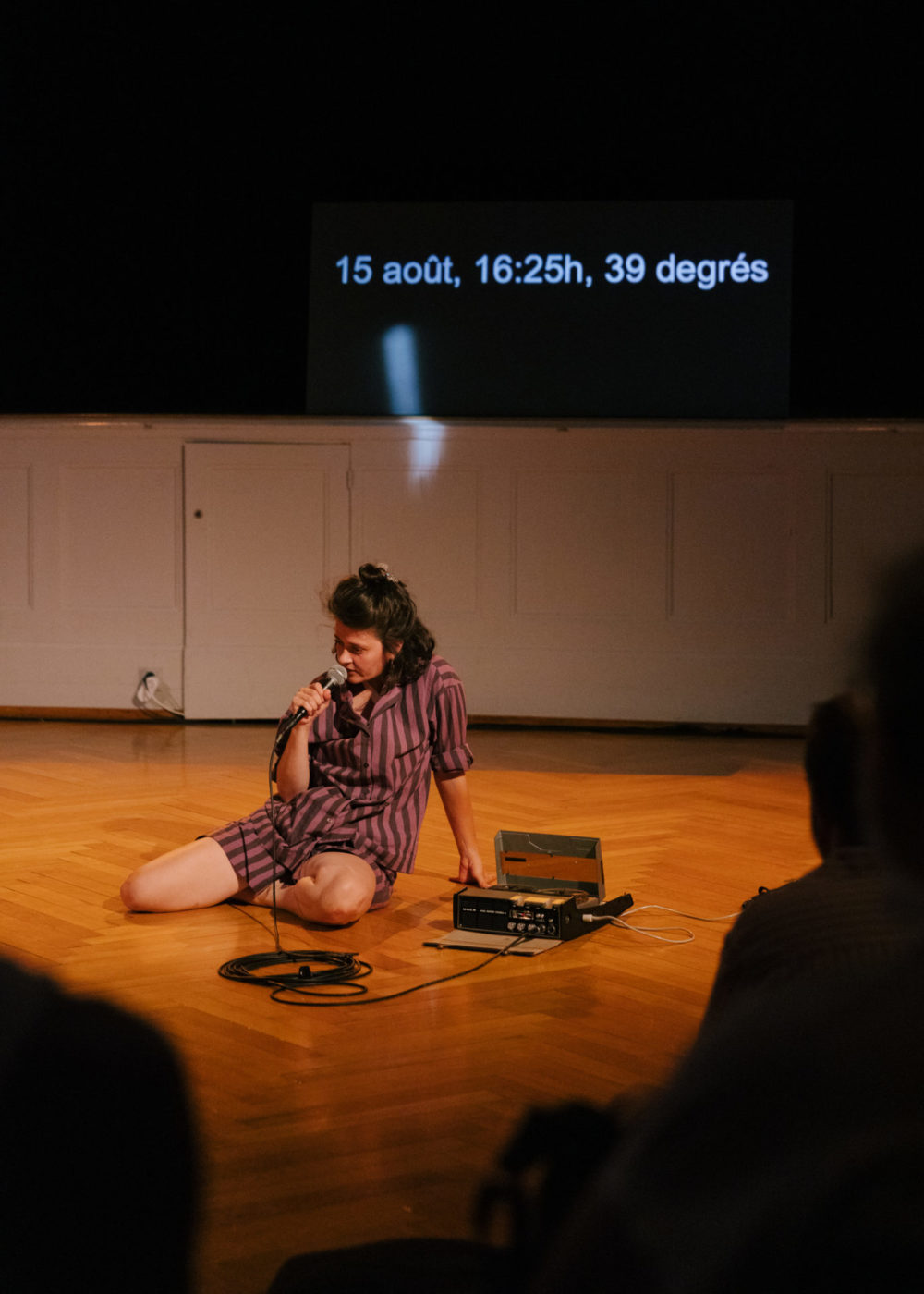 L'artiste Annina Polivka se tient assise par terre, devant la scène. Elle a un micro dans sa main dans lequel elle parle.