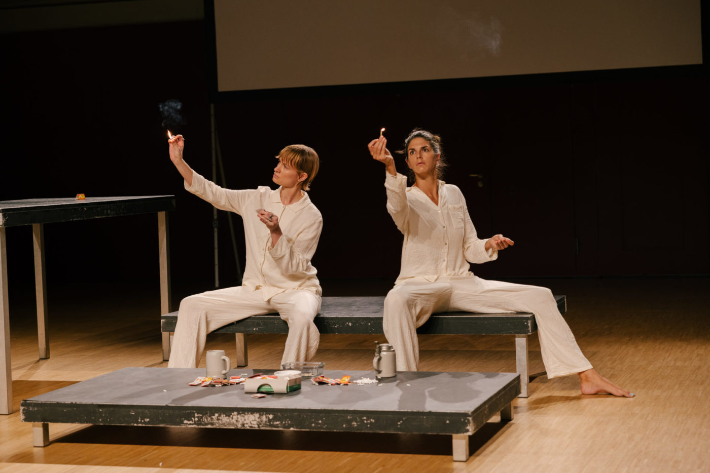 Les deux artistes Flavia Papadaniel et Diane Dormet sont sur scène, assises sur un banc. Elles portent un pyjama. Chacune tient de la main droite une allumette allumée en la regardant fixement.